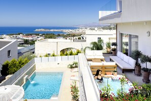 A rooftop pool with a view of the sea, a white outdoor seating area, and a modern building with large windows.