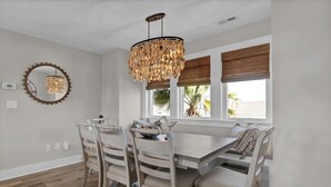 A dining area with a wooden table and chairs, a chandelier, and a view of palm trees through the windows. - Ocean & Sound Views, Elevator, Close to Beach & Dining! (Wrightsville Beach)