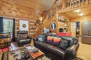 A cabin interior with a wooden interior, a leather sofa, a wooden coffee table, a fireplace, and a kitchen area. - 2BR Getaway with Hot Tub, Firepit & Outdoor TV Fun! (Gatlinburg)