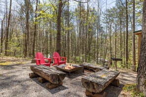 A forested campsite with a fire pit, two red Adirondack chairs, and a wooden table. - 2BR Getaway with Hot Tub, Firepit & Outdoor TV Fun! (Gatlinburg)