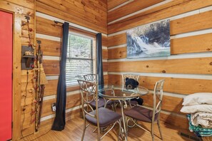 A cabin interior with a round glass table, four chairs, a bear decoration, a painting of a waterfall, and a red door. - 2BR Getaway with Hot Tub, Firepit & Outdoor TV Fun! (Gatlinburg)