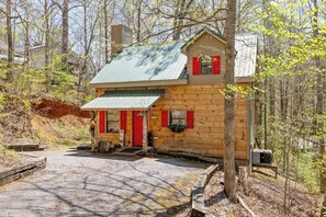 A wooden cabin with a red door and windows, surrounded by trees and a gravel driveway. - 2BR Getaway with Hot Tub, Firepit & Outdoor TV Fun! (Gatlinburg)