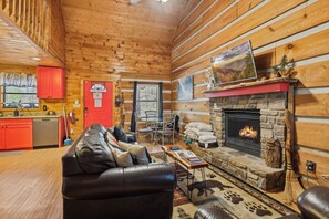 A cozy cabin interior with a stone fireplace, wooden walls, a leather sofa, and a dining area. - 2BR Getaway with Hot Tub, Firepit & Outdoor TV Fun! (Gatlinburg)