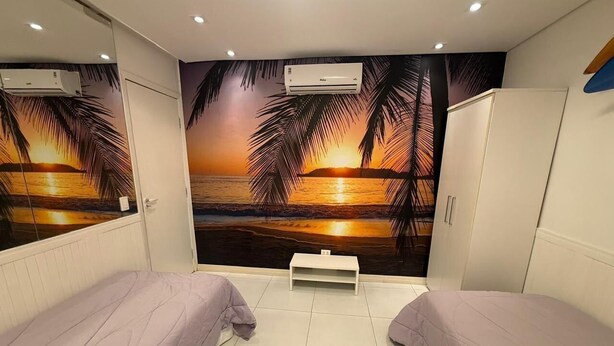Une chambre avec deux lits, une petite table blanche et une fresque murale représentant un coucher de soleil sur l’eau.