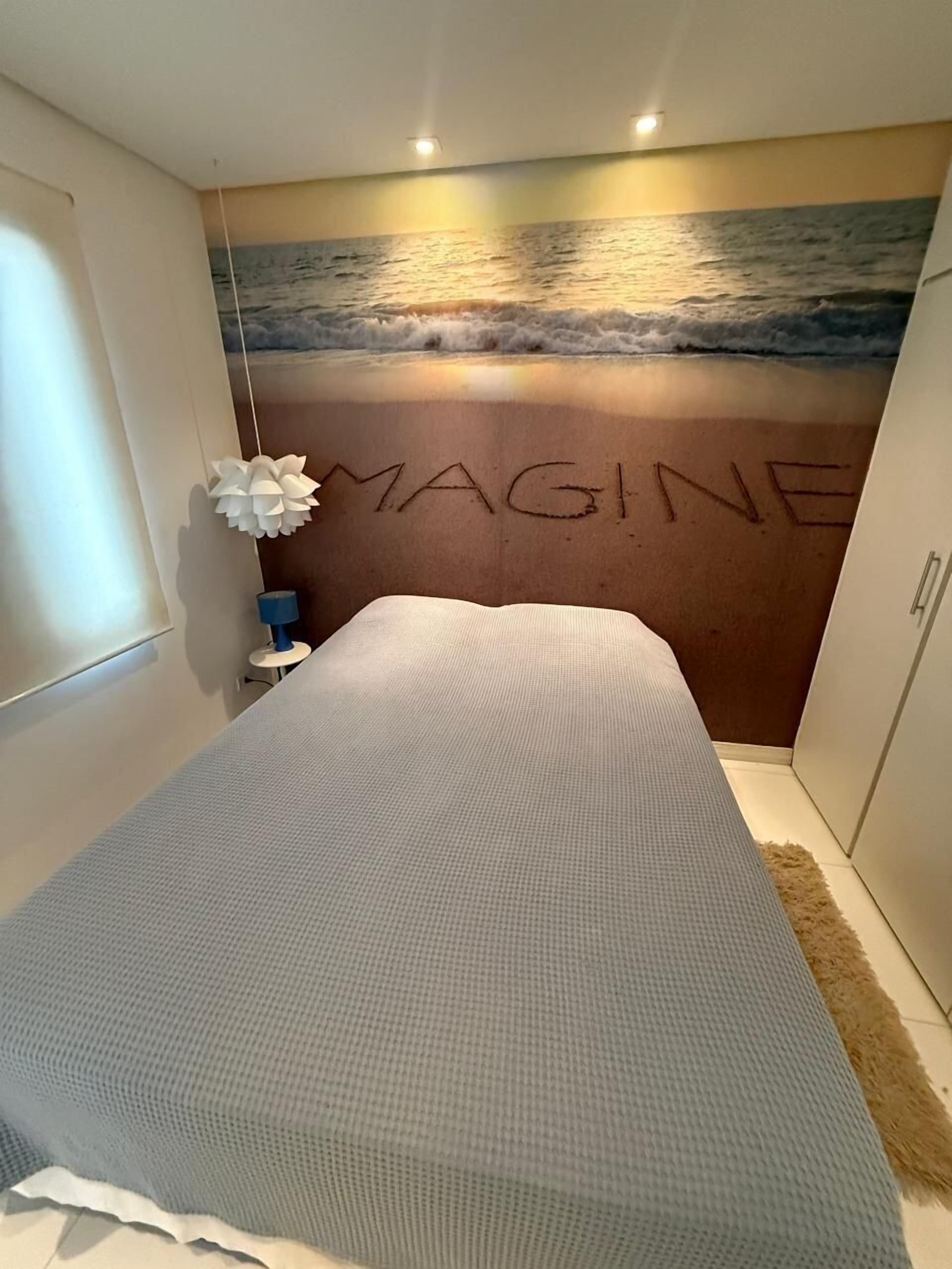 A bedroom with a bed, a wall mural of a beach, and the word "MAGINE" written on the wall.