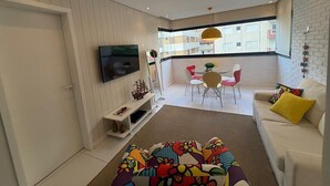 Un salon moderne doté d’un téléviseur à écran plat, d’un canapé agrémenté de coussins colorés, d’un coin repas avec une table et des chaises, et d’un balcon offrant une vue sur les immeubles.