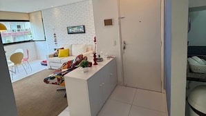 Un salon moderne avec un canapé, une table basse et un téléviseur fixé sur un mur en briques blanches. Une porte donne accès à une autre pièce et une fenêtre apporte de la lumière naturelle.