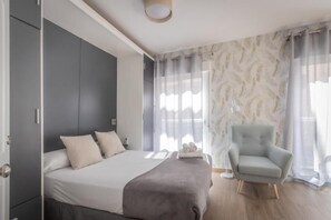 Um quarto moderno com uma cama, uma poltrona cinzenta e uma janela grande com cortinas leves.