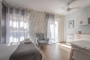 Quarto de hotel moderno com cama, cadeira, escrivaninha e cozinha compacta.