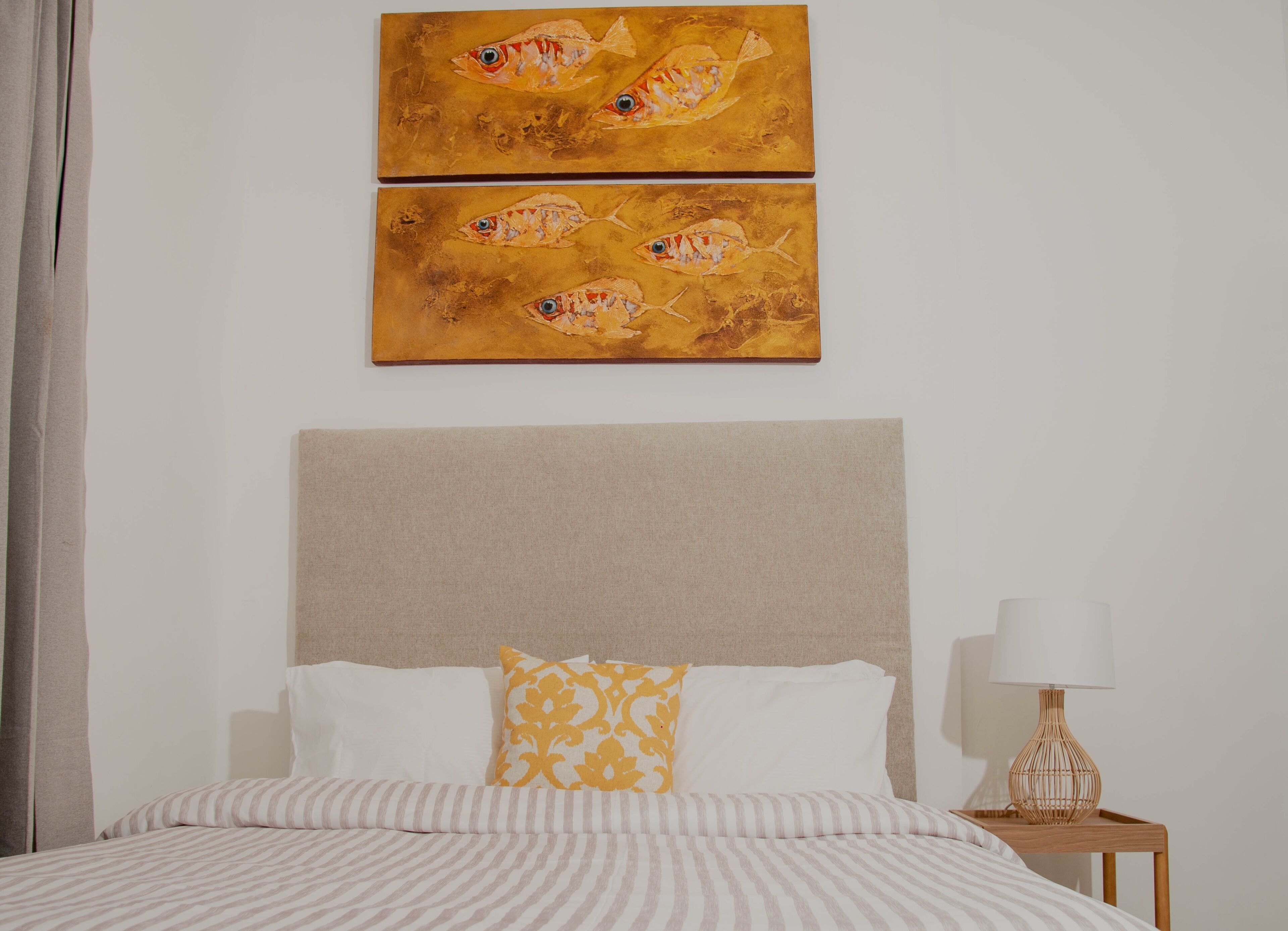 A bed with striped bedding, a headboard, two paintings of fish, a lamp, and a curtain.