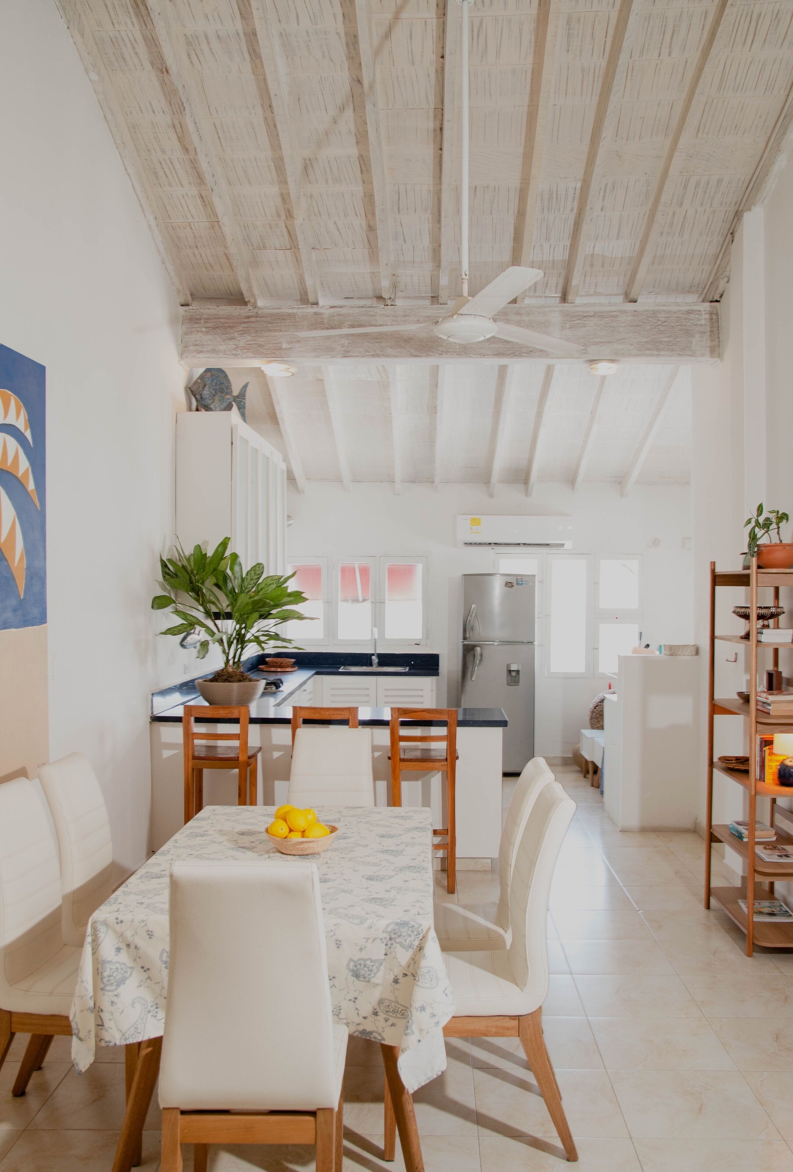 A modern kitchen with white cabinets, a built-in oven, and a refrigerator. There is a dining area with a table set for two, chairs, and a potted plant.