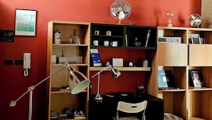 A room with red walls, wooden bookshelves, a desk with a chair, a desk lamp, a wall-mounted fan, and a framed picture. - Cozy and quiet apartment in ideal location in Reggio Cal. (Reggio Calabria)