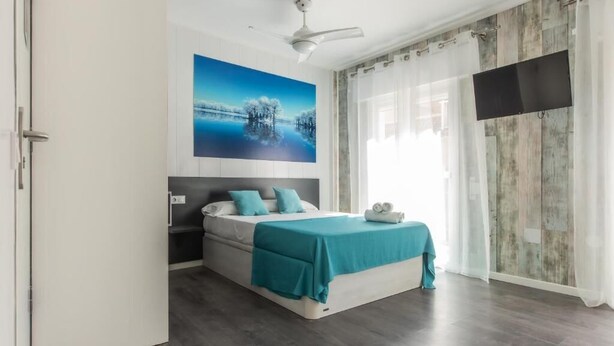 Un dormitorio moderno con cama, ventilador de techo, televisor montado y un ventanal con cortinas.