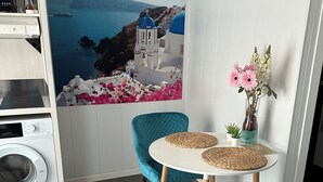 Una habitación compacta con lavadora, una mesa con dos sillas, un florero con flores y un cuadro colgado en la pared que representa una escena costera.