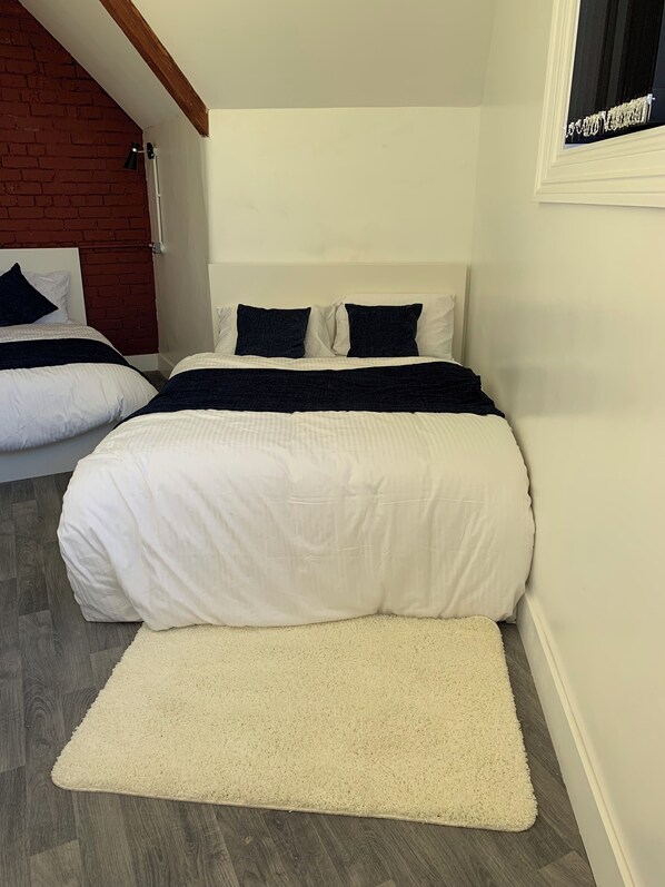 A bedroom with two beds, white bedding, a white rug, a wooden ceiling, and a brick wall. - Rahman Hostel - Manchester (Manchester)