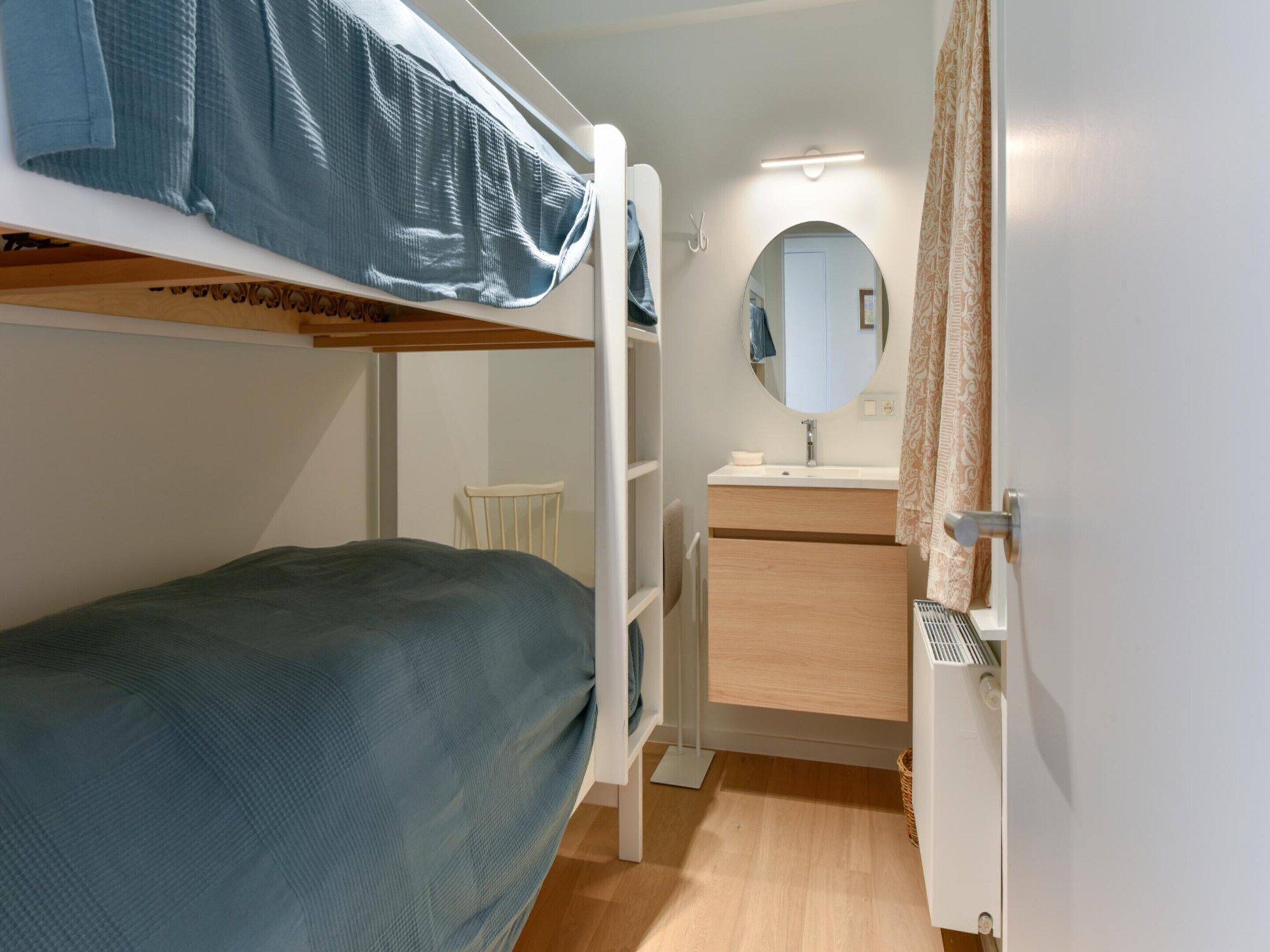 A bunk bed with a blue mattress, a small white chair, a wooden cabinet with a mirror, and a bathroom with a sink and shower.