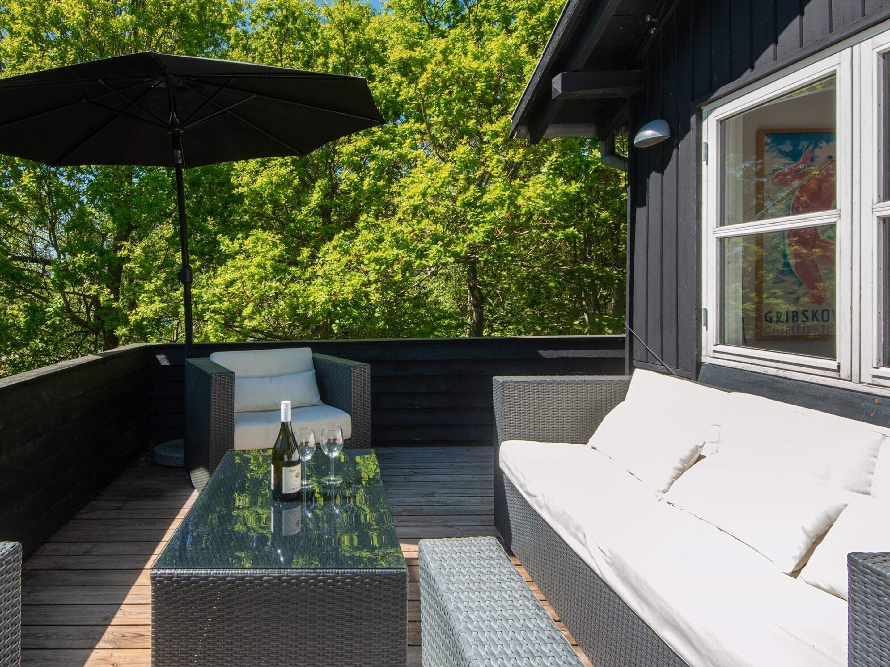 Eine Terrasse mit Korbmöbeln, einem Glastisch mit Flasche und Gläsern sowie einem Sonnenschirm.
