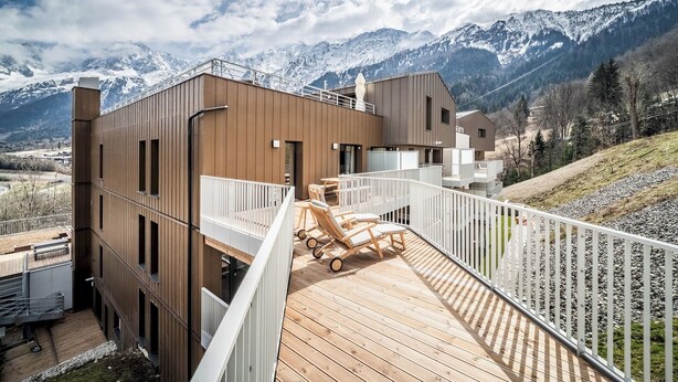 Een modern gebouw met een houten terras en loungestoelen met uitzicht op een bergachtig landschap.