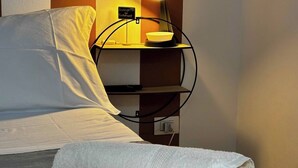 Een net opgemaakt bed met een opgerolde witte handdoek, een bedlamp en een klein tafeltje met een kom en een bordje.