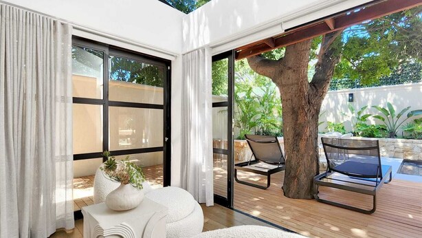 A modern living room with a large glass door leading to a wooden deck and outdoor seating area. - Botany House Boutique Retreat (Cape Town)