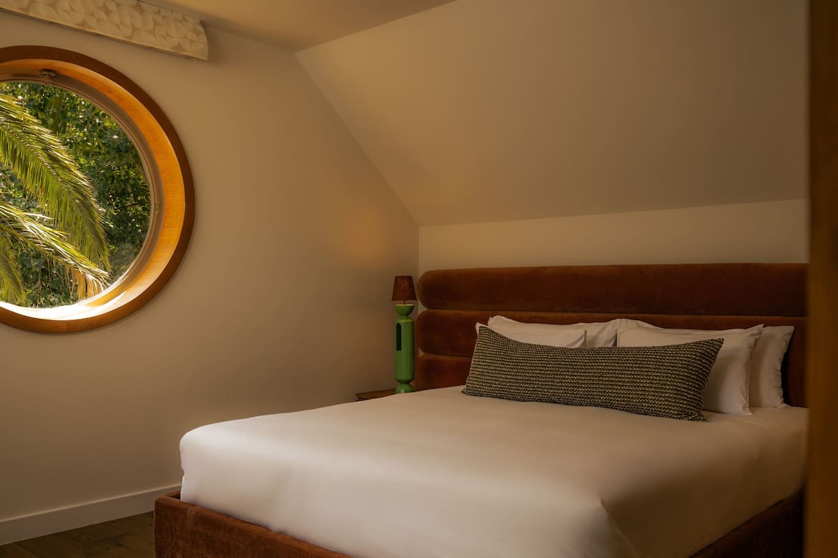 Quarto com cama, janela redonda e vista para área verde.
