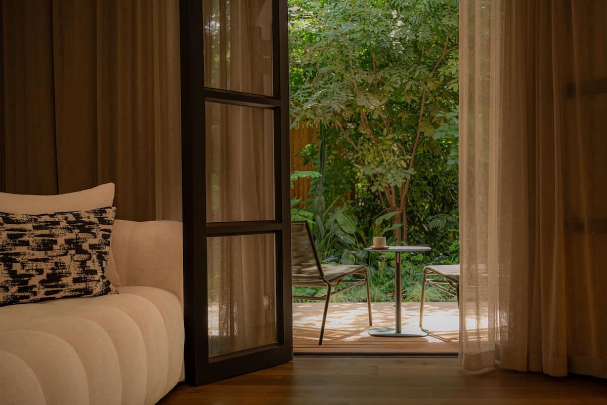 Quarto com cama, cadeira e mesa com uma xícara. Há uma porta que dá para área externa com plantas e deck de madeira.
