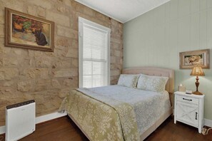 A bedroom with a stone wall, a bed with a floral bedspread, a nightstand with a lamp, and a radiator.