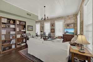 A living room with a white sofa, a wooden bookshelf, a dining table with chairs, a chandelier, and a television.