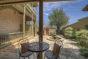Une cour avec un sol en pierre, deux tables en métal avec des chaises, une petite fontaine et un bâtiment doté d’un balcon.
