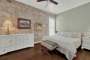 A bedroom with a stone wall, a bed with white bedding, a nightstand, a lamp, and a ceiling fan.