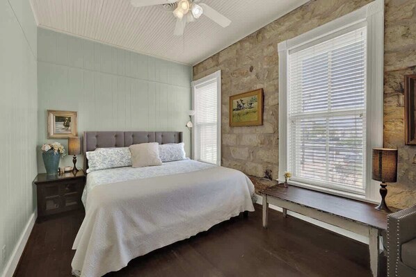 A bedroom with a bed, a nightstand, a bench, a ceiling fan, and stone walls. - Charming Balcony Suite Overlooking Main Street! (Fredericksburg)