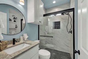Ein modernes Badezimmer mit Waschbecken aus Granit, einer weißen Toilette, einer Dusche mit Glaswand und einem Spiegel mit dekorativer Pflanze.
