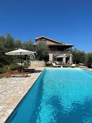 Une piscine entourée d’une terrasse en pierre, agrémentée de chaises longues et de parasols, et une maison avec un balcon.