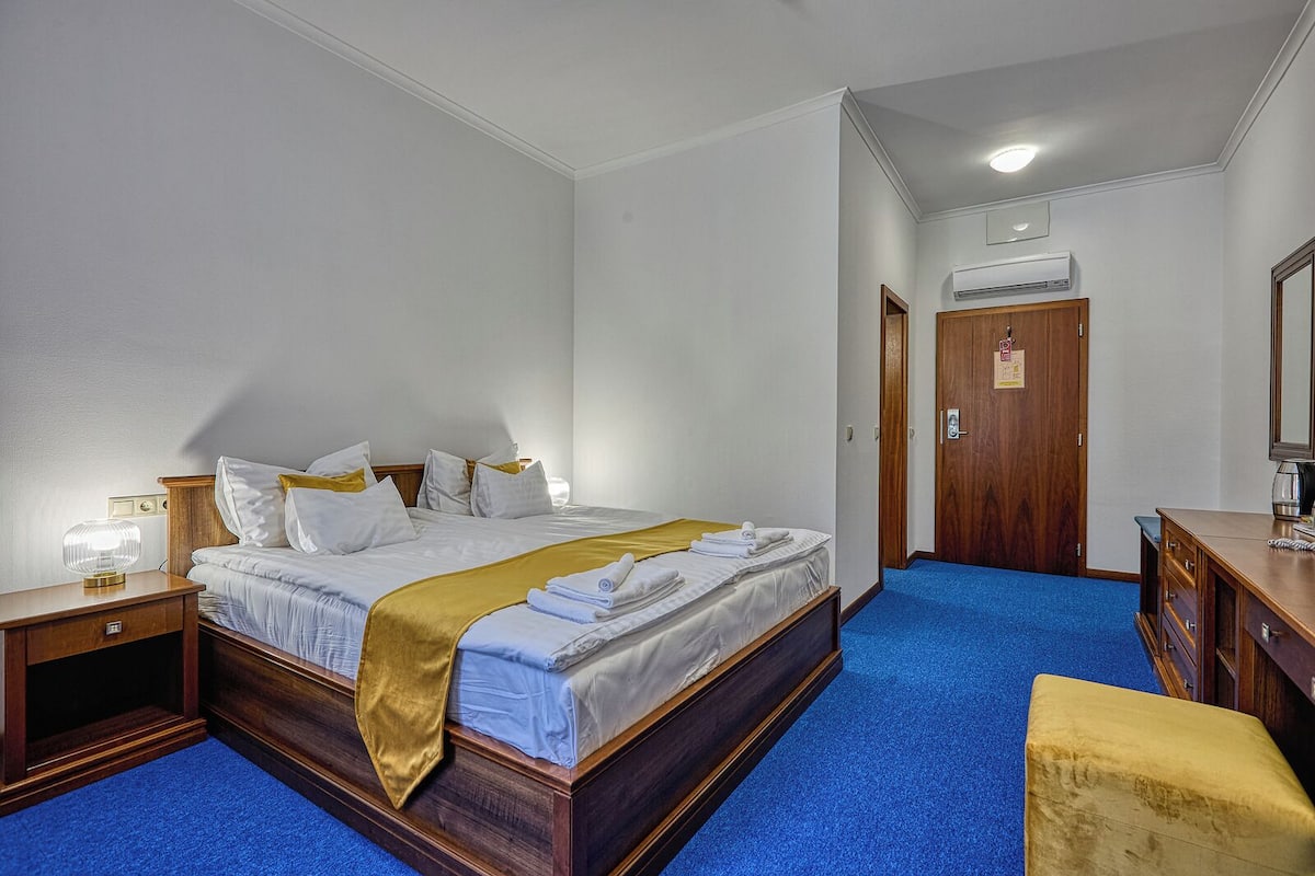 A hotel room with a large bed, wooden headboard, a bedside table with a lamp, a wardrobe, and a desk with a chair.