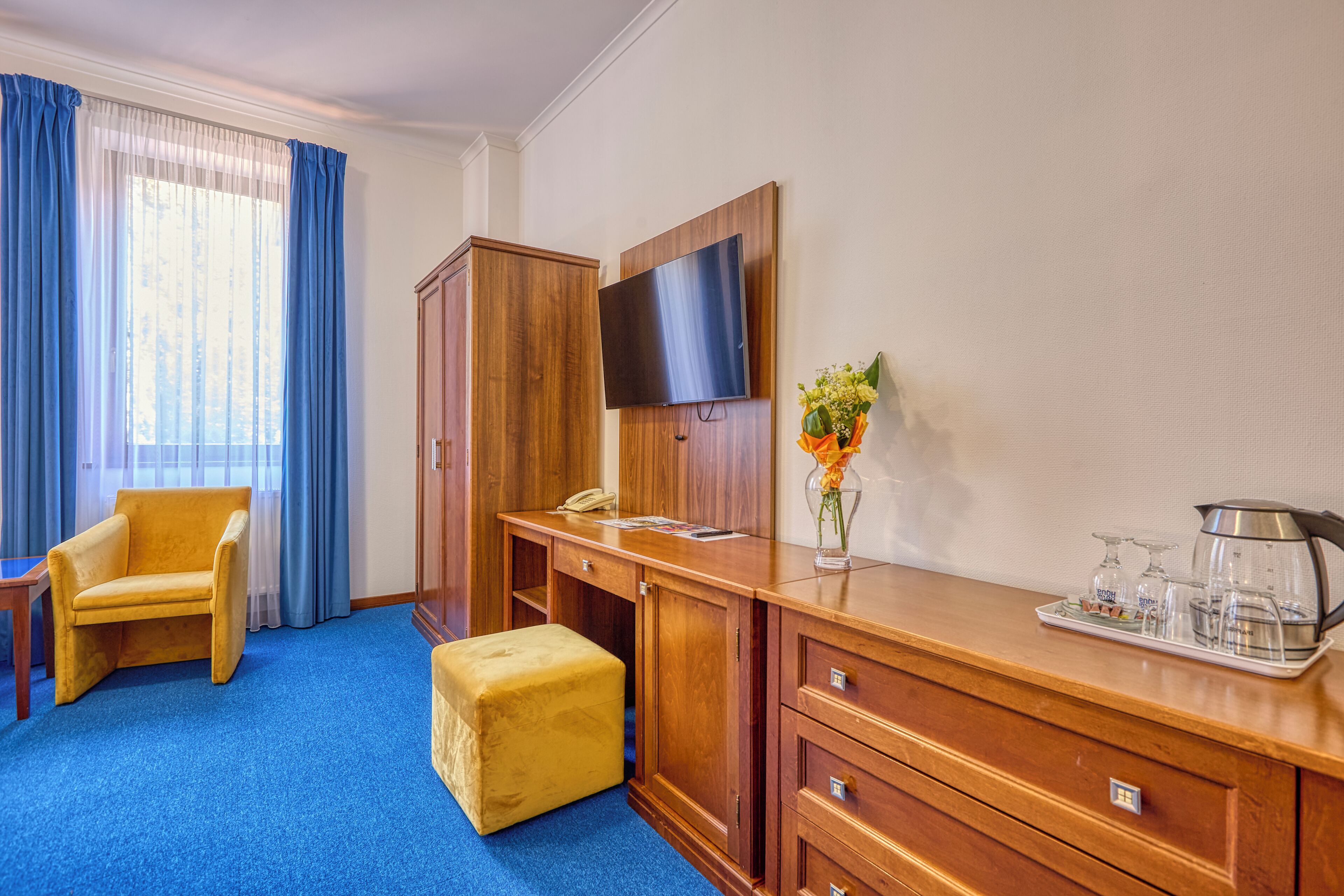 A hotel room with a wooden desk, a television, a telephone, a vase with flowers, and a tray with glasses and a kettle.