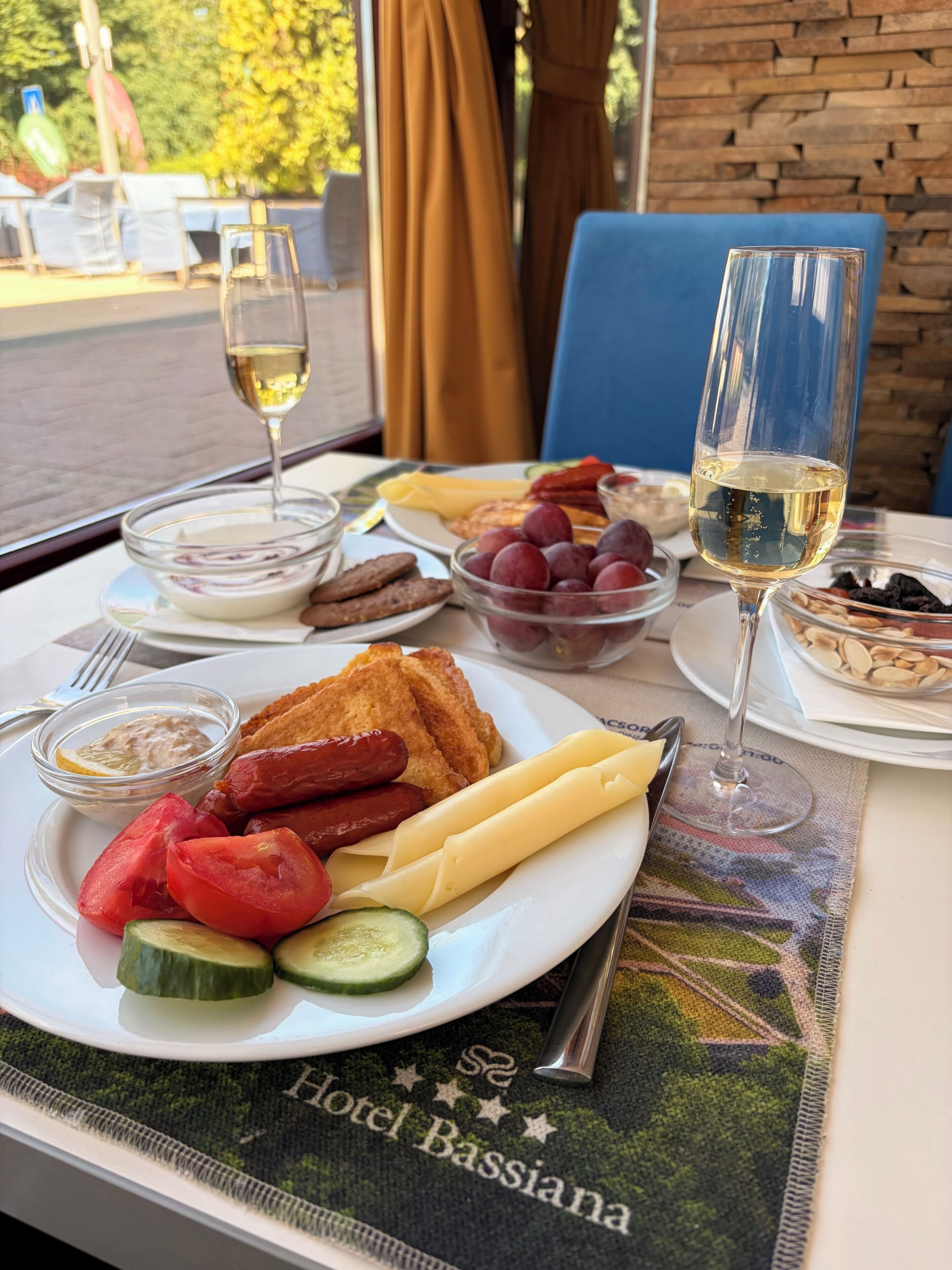 Uma mesa de jantar com um prato de comida, um copo de vinho branco e um menu com o texto 'Hotel Bassiana'.