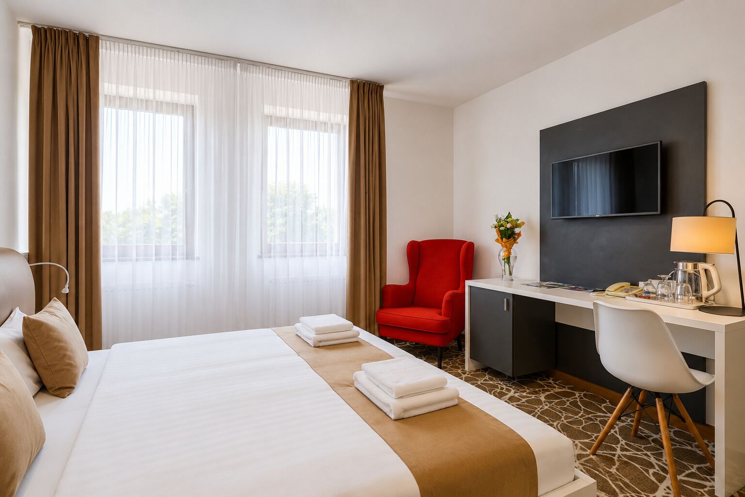A hotel room with a bed, a desk, a red chair, a television, and a window with curtains.