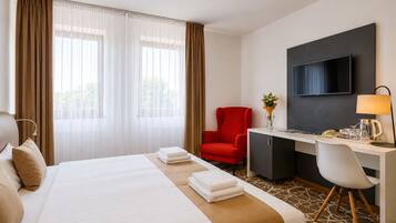 A hotel room with a bed, a desk, a red chair, a television, and a window with curtains.