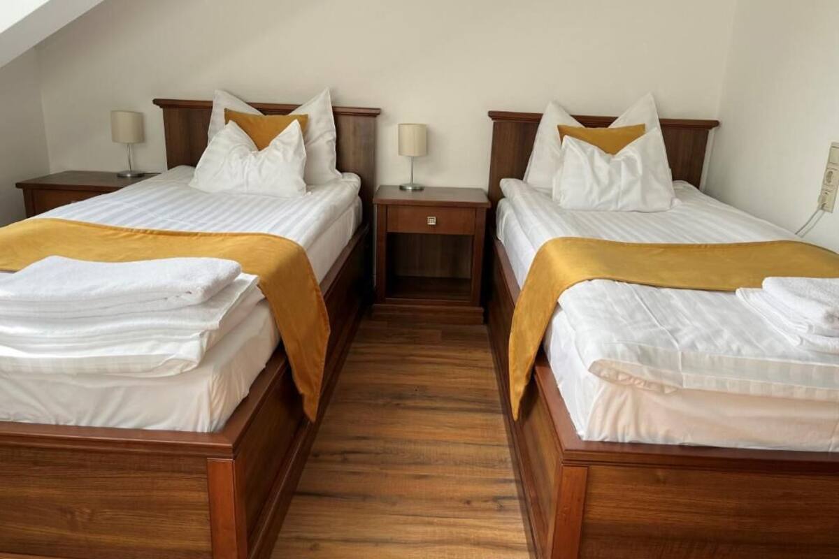 A hotel room with two wooden beds, each with a white mattress and a yellow throw blanket. There are bedside tables with lamps and a small drawer.