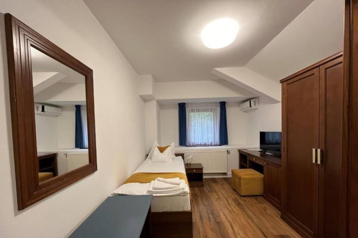 A small, well-lit room with a bed, a bench, a desk, and a wardrobe.