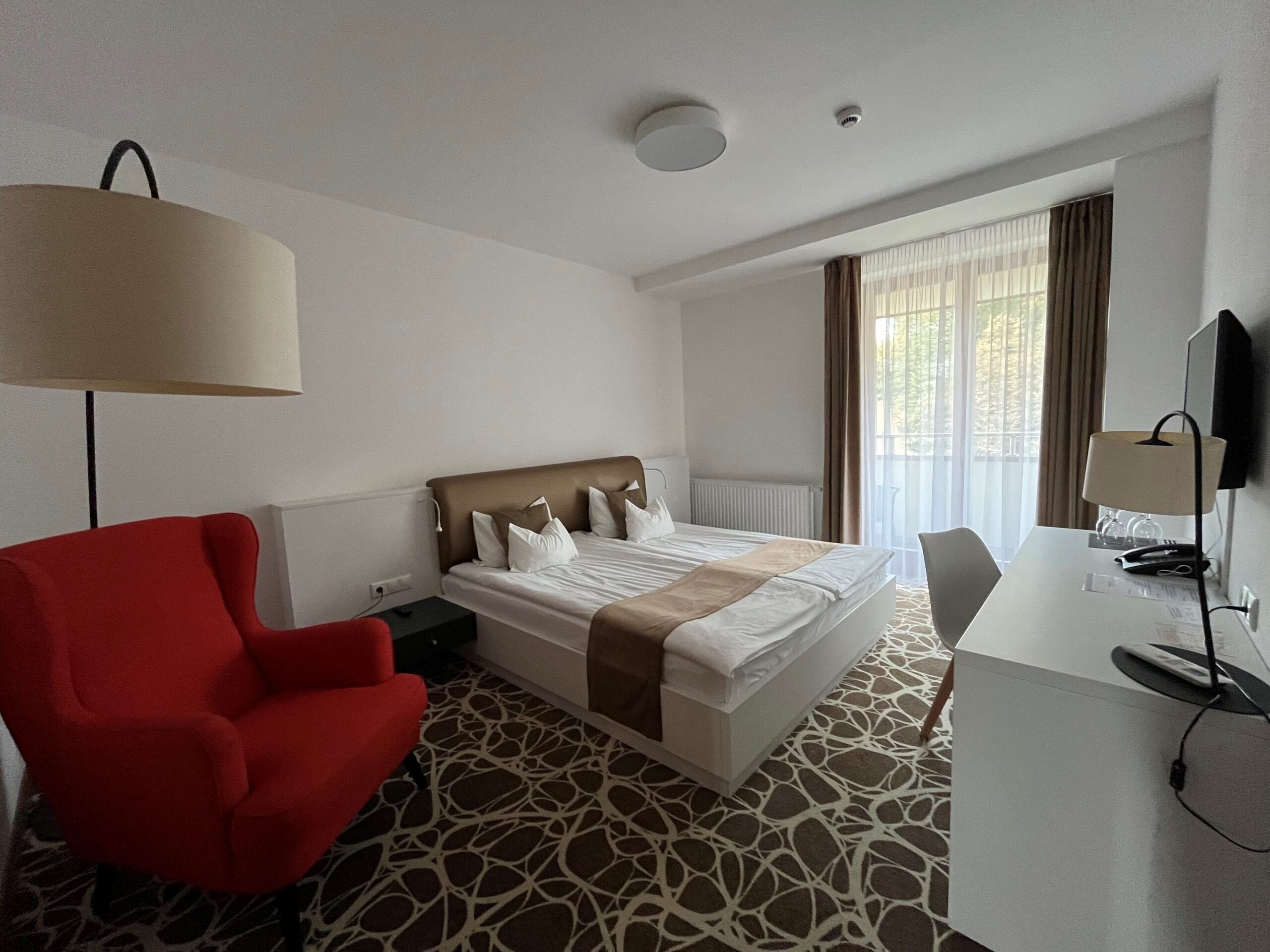 A hotel room with a bed, a desk, a chair, a lamp, and a window with curtains.