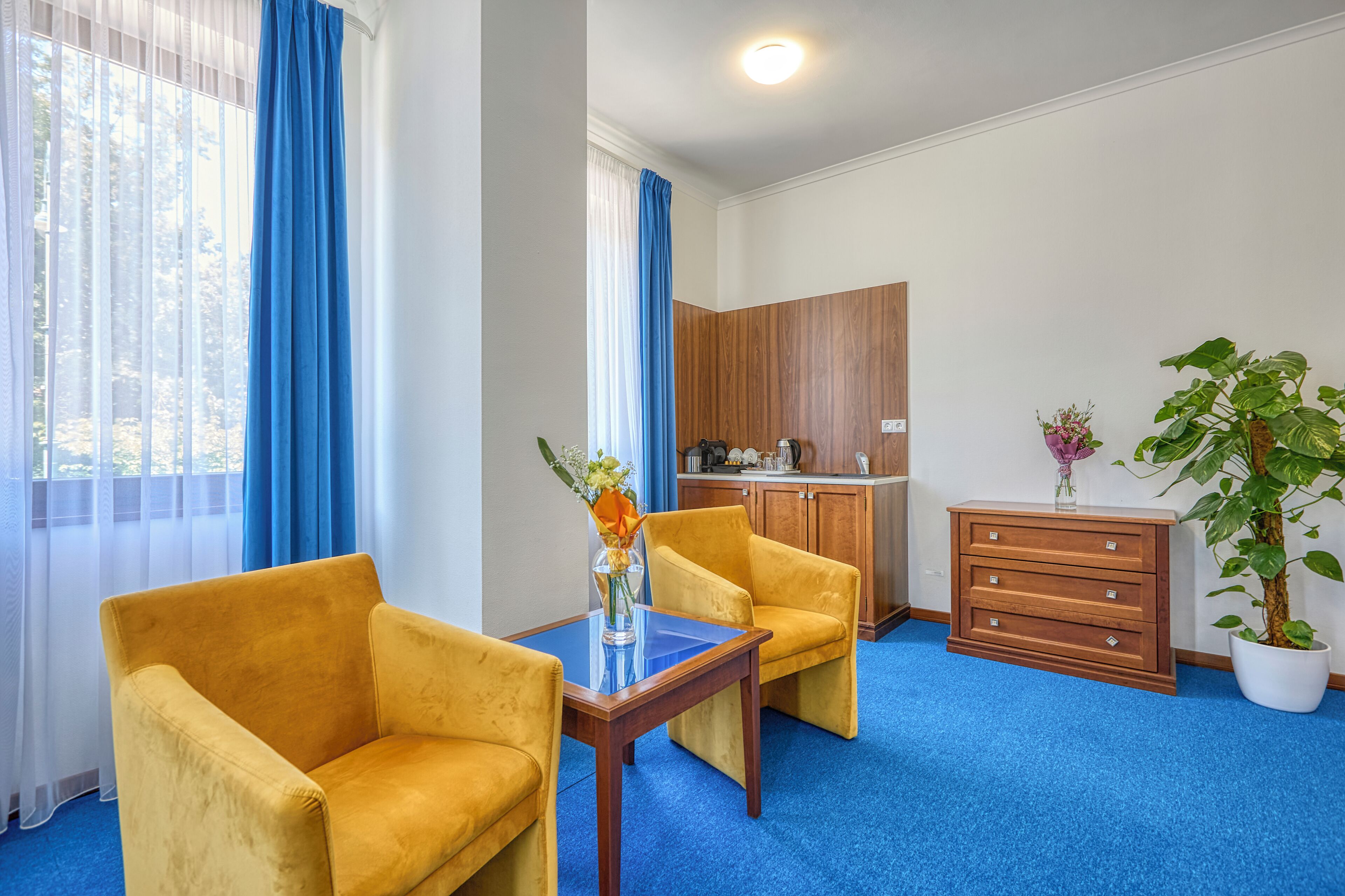 A hotel room with two yellow armchairs, a wooden cabinet, a small table with a vase, and a potted plant.
