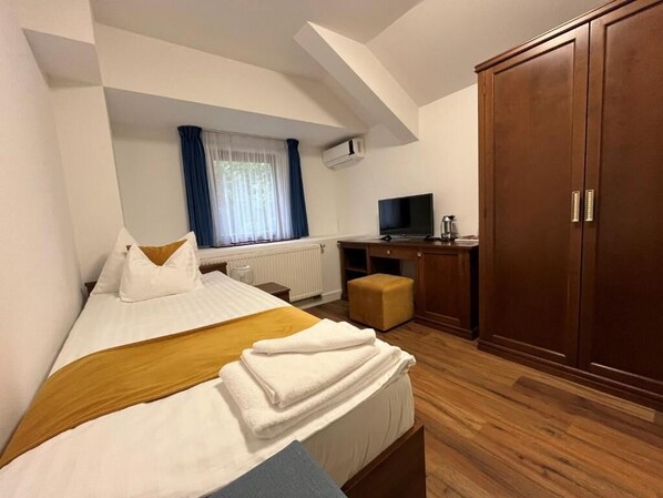 A hotel room with a bed, a desk with a television, a wardrobe, and a window with curtains. - Hotel Bassiana (Sárvár)
