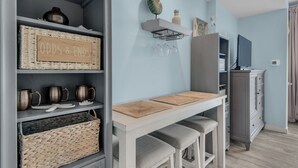 A modern kitchen with a light blue wall, a wooden table with stools, a built-in shelf with kitchenware, and a grey cabinet. - Quaint Efficiency Suite-Sleeps 4-Resort Amenities (Miramar Beach)