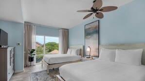 A hotel room with two beds, a ceiling fan, a large window with curtains, a TV, and a painting on the wall. - Quaint Efficiency Suite-Sleeps 4-Resort Amenities (Miramar Beach)