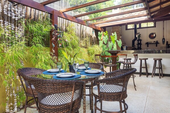 A covered outdoor dining area with wicker chairs and a glass table set for four. - Bz62 Centro Casa 5 Suítes e área gourmet (Buzios)
