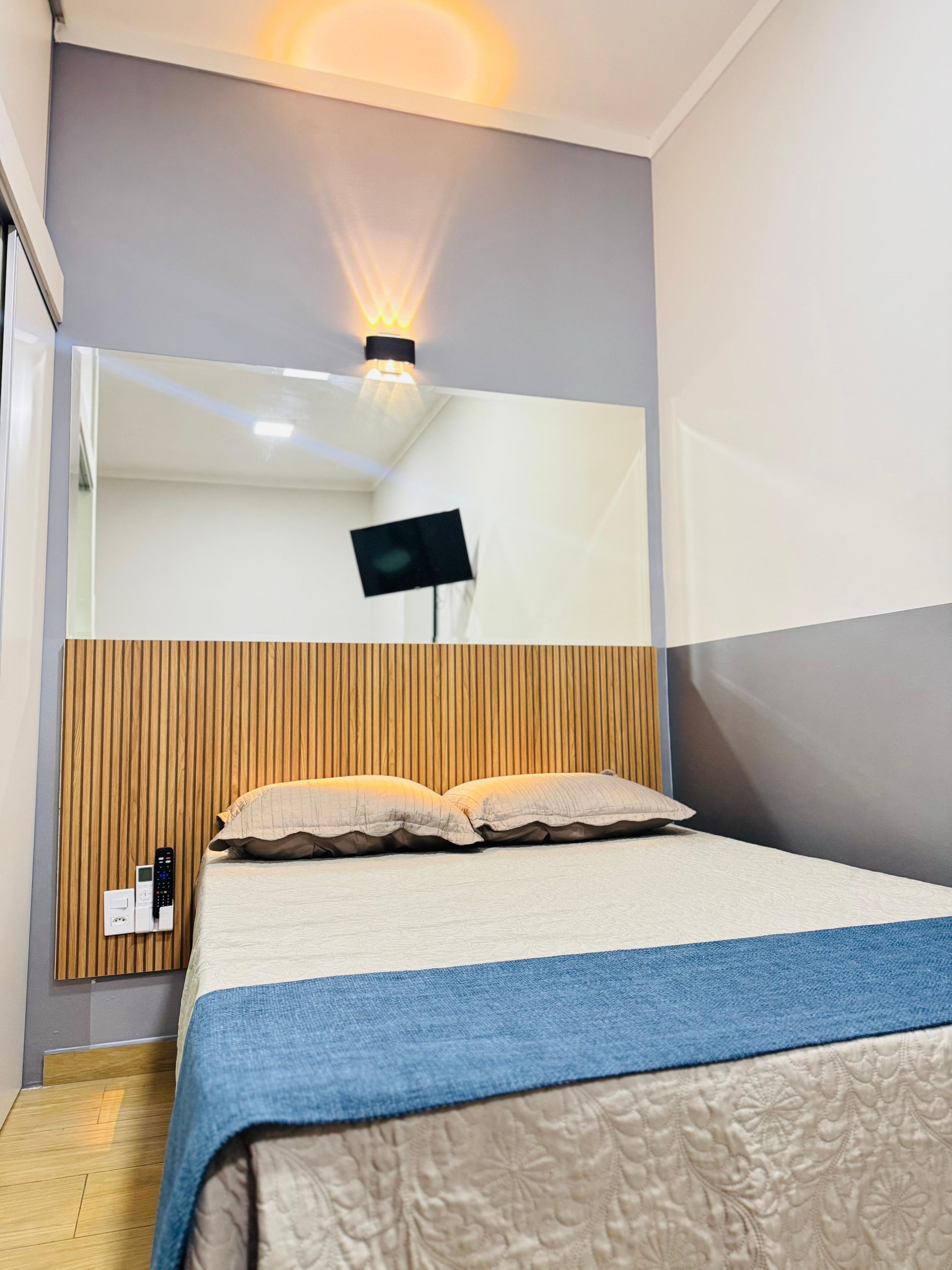 Un dormitorio moderno con una cama, un televisor montado y una mesita de noche con un control remoto.