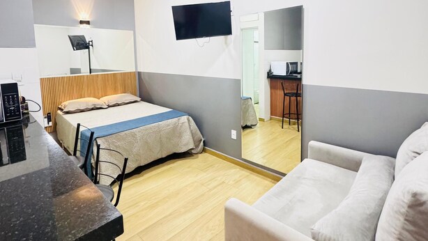 A modern hotel room with a bed, a sofa, a desk, and a flat-screen TV mounted on the wall. - Residencial Aurelio Souza (Pouso Alegre)