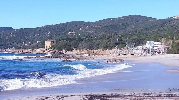 Una playa con olas, un edificio y colinas al fondo.