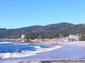 A beach with waves, a building, and hills in the background.